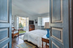 A bedroom with a large bed, a wooden dresser, a chair, and a table with a lamp. There is a window and a door leading to an outdoor area.
