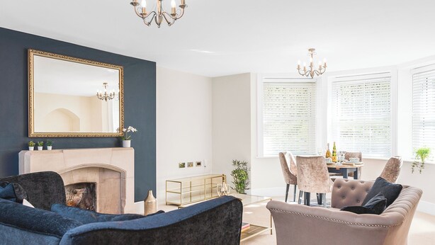 Ein Wohnzimmer mit Kamin, zwei Sesseln, einer Couch, einem Esstisch mit Stühlen und einem großen Spiegel an der Wand.