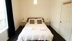 A neatly made bed with white linens and two pillows, two bedside tables with lamps, a window with black curtains, and a white door with a coat rack. - Gloucester Apartment - UK37842 (Bristol)