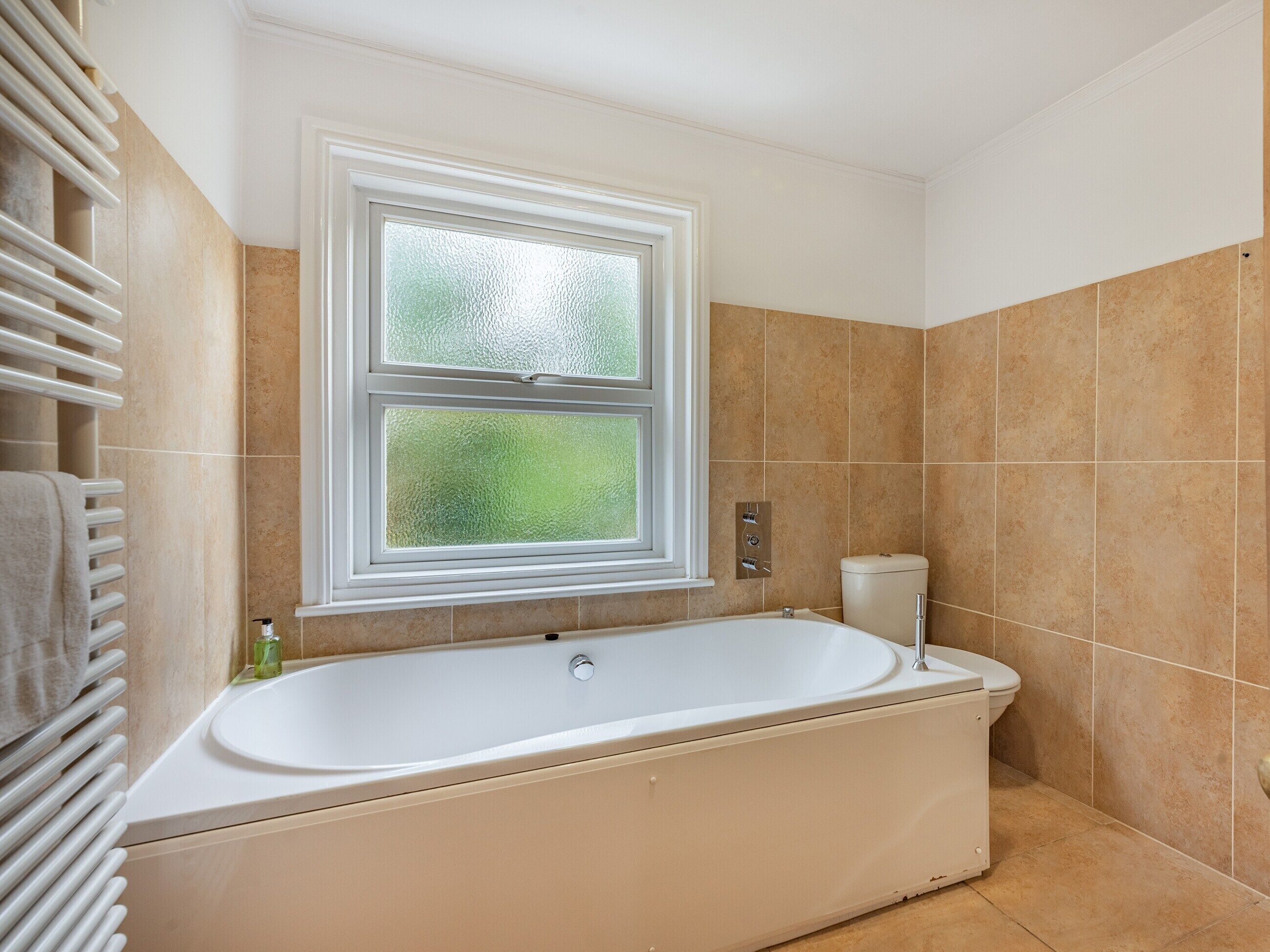 A bathroom with a freestanding bathtub, a window with frosted glass, and beige tiled walls.