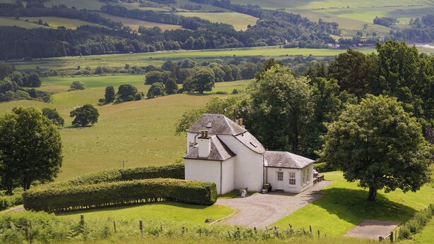 A white building with a sloped roof, surrounded by green fields and trees, with a clear sky above. - Castle Peroch-UK5698 (By Dunkeld, Pitlochry)