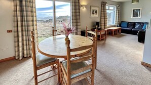 A room with a round wooden dining table, chairs, a sofa, and a view of the outdoors. - Orchid Cottage - UK5866 (Clachtoll, near Lochinver)