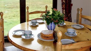 A round wooden table with plates, cups, a cake, and a vase with flowers. - Orchid Cottage - UK5866 (Clachtoll, near Lochinver)