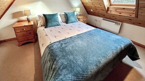 A bedroom with a sloped ceiling, a bed with a blue blanket, two bedside tables with lamps, and a window with a view. - Orchid Cottage - UK5866 (Clachtoll, near Lochinver)