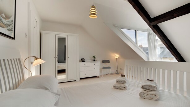 Un dormitorio con techo inclinado, una cama con ropa blanca, un aparador, una silla y una lámpara de pie.
