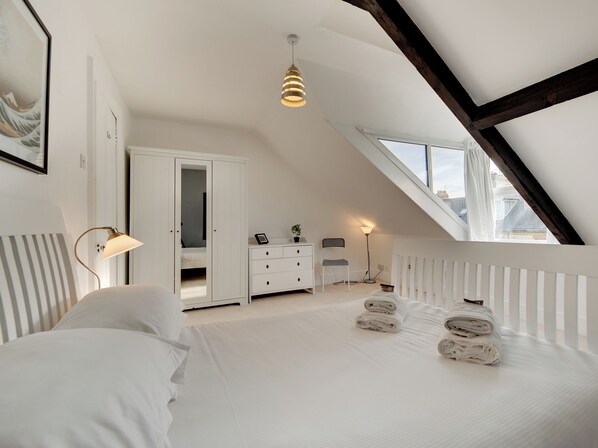 Ein Schlafzimmer mit schrägverlaufender Decke, einem Bett mit weißen Leinen, einem Schminktisch, einem Stuhl und einer Stehlampe.