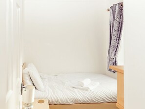 Un petit lit bien fait, avec du linge de lit blanc et une serviette enroulée, une table de chevet en bois et un mur blanc avec une fenêtre.