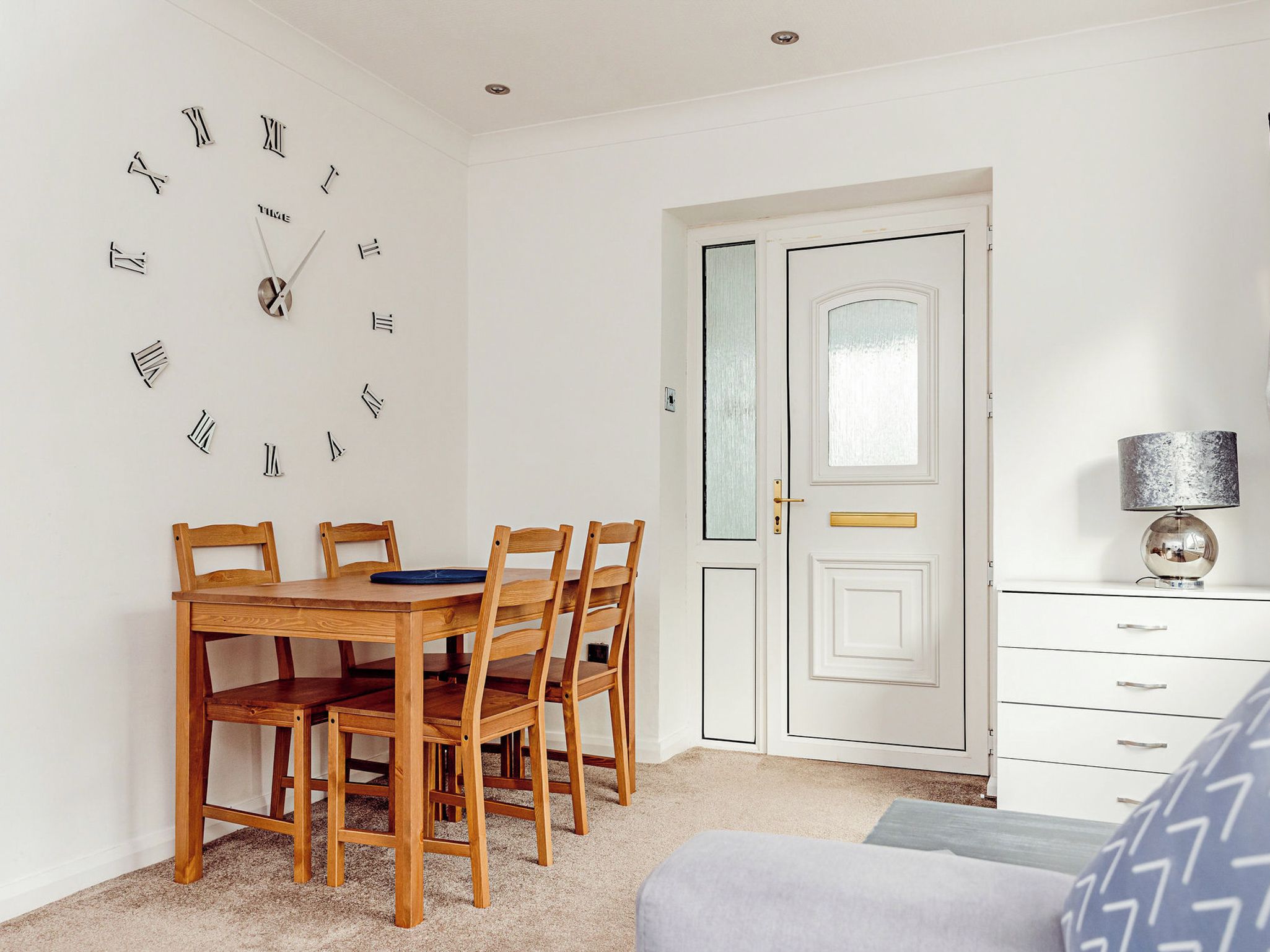 Un coin repas avec une table et des chaises en bois, une horloge murale, une porte blanche avec des panneaux en verre dépoli et une lampe posée sur une commode.