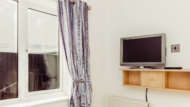 Ett rum med en TV på en hylla, ett fönster med gardin och ett element.