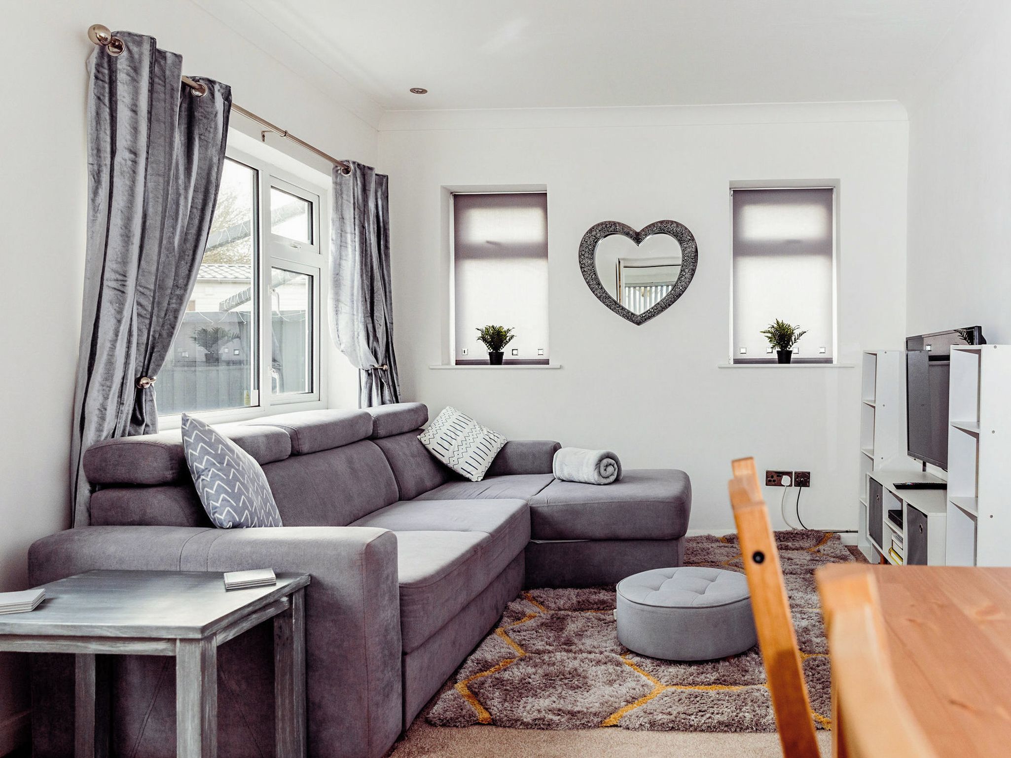Ein modernes Wohnzimmer mit einem grauen Ecksofa, einem runden Hocker, einem kleinen Tisch und einer TV-Konsole mit einem Flachbildfernseher.