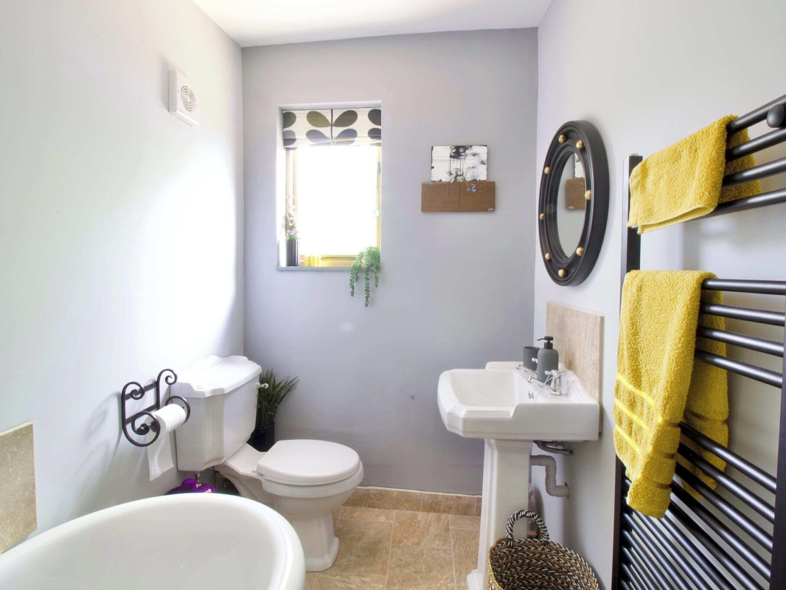 A bathroom with a toilet, sink, and bathtub. There is a window with a view of greenery outside, a towel rack with yellow towels, and a decorative mirror.