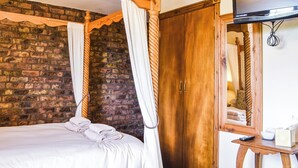 Una cama con dosel con sábanas blancas, un armario de madera, un televisor sobre un atril de madera y una pared de ladrillo.