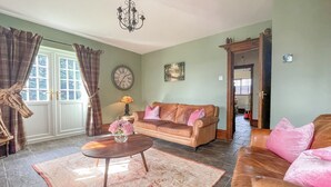 Un salón con un sofá de cuero marrón, una mesa de centro con un jarrón de flores, un reloj en la pared y una puerta que da a otra habitación.
