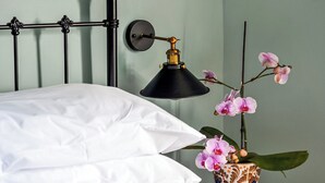 Una cama bien hecha con ropa blanca, una lámpara negra fijada a la pared y un jarrón floral con orquídeas sobre una mesita de noche.