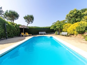 A rectangular swimming pool with a white edge, surrounded by a paved deck. There are two yellow lounge chairs and a white table with a chair. The area is bordered by well-maintained hedges and trees, with a clear blue sky above. - Villa in Spain for 20 people, near beaches (Caldes de Malavella)