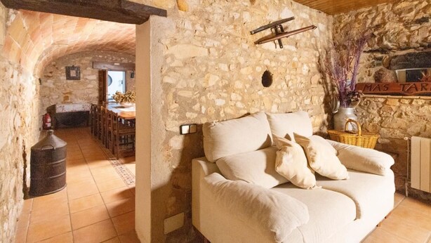 A stone-walled room with a wooden ceiling, a sofa with cushions, a fireplace, and a dining area visible through an archway.
