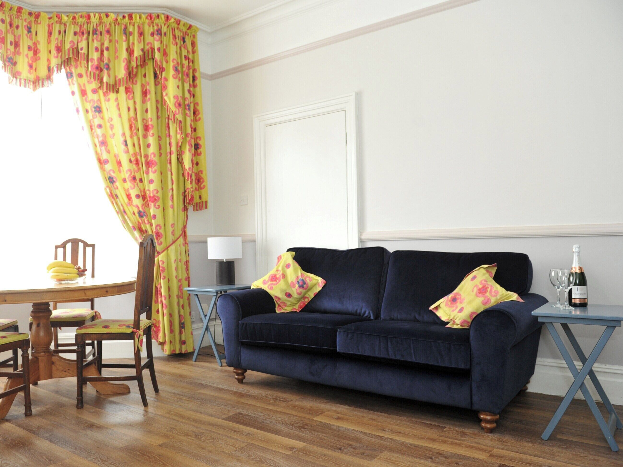 A living room with a dark blue sofa, two floral cushions, a wooden dining table with chairs, a small side table with a lamp, and a bottle of champagne on a stand.