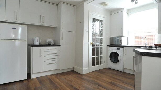 A modern kitchen with white cabinetry, a built-in washing machine, and a large window providing natural light. - 1 bedroom accommodation in Cheltenham (Cheltenham)