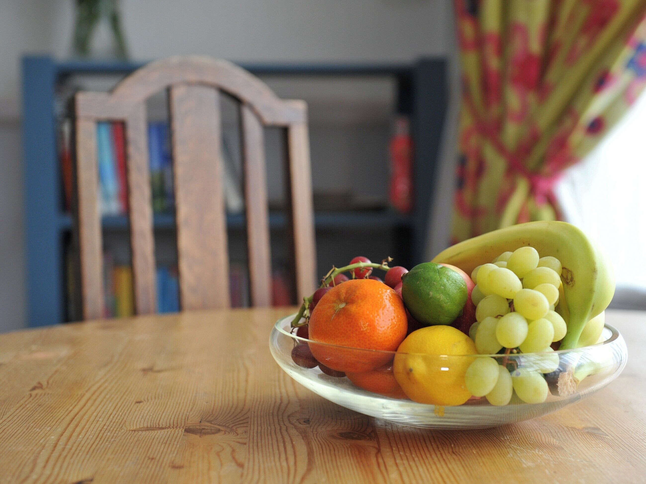 A wooden table with a glass bowl containing an orange, a lime, a banana, and grapes.