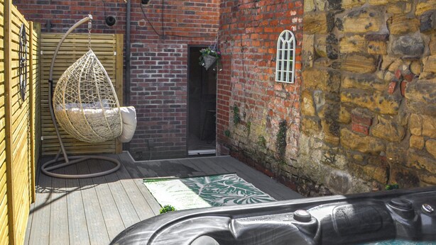 A wooden deck with a hot tub, a hanging chair, and a brick wall. - 2 bedroom accommodation in Morpeth (Morpeth)
