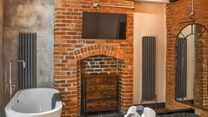 A bathroom with a freestanding bathtub, a brick fireplace, a wooden dresser, and a mirror. - 2 bedroom accommodation in Morpeth (Morpeth)