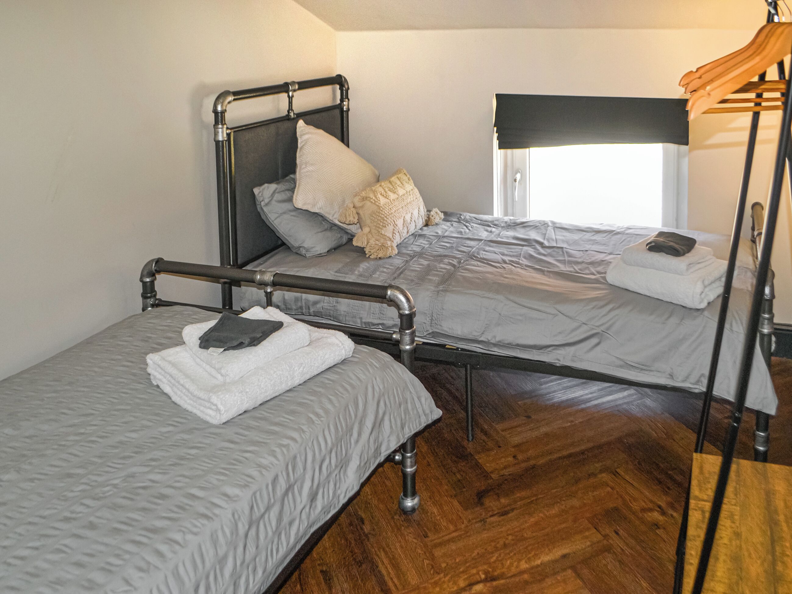 Una habitación con dos camas, cada una con estructura metálica y ropa de cama gris. Se observa una ventana con persiana negra y un perchero de madera.