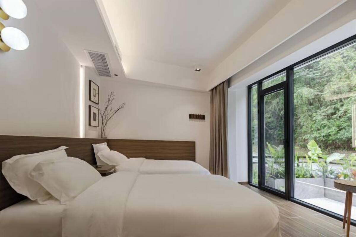 A modern hotel room with a large bed, a small table, and a view of greenery through large windows.
