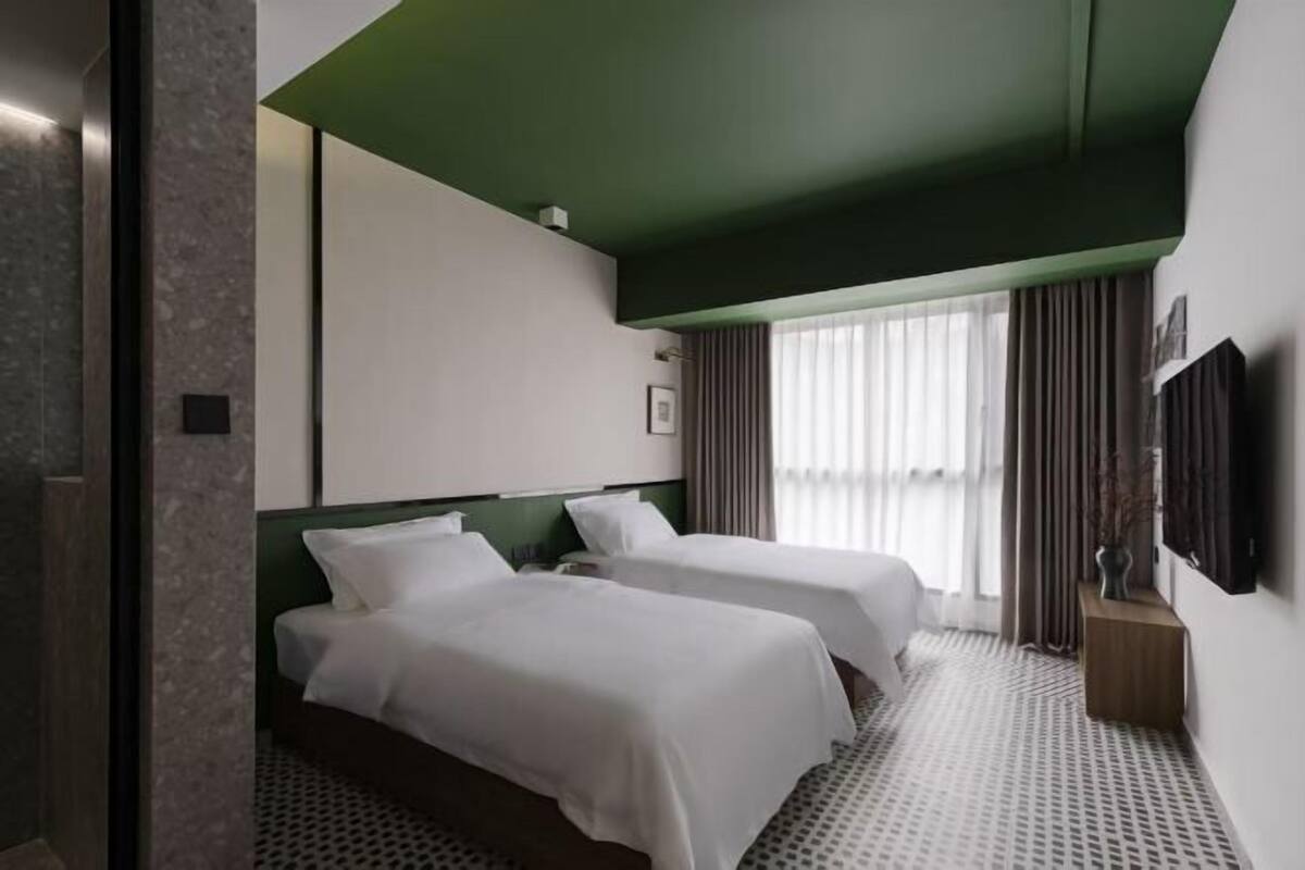 A hotel room with two beds, a green accent wall, a large window with curtains, and a mounted TV.