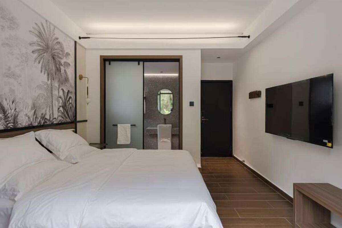 A hotel room with a large bed, a flat-screen TV, a painting on the wall, and a bathroom visible through a glass door.
