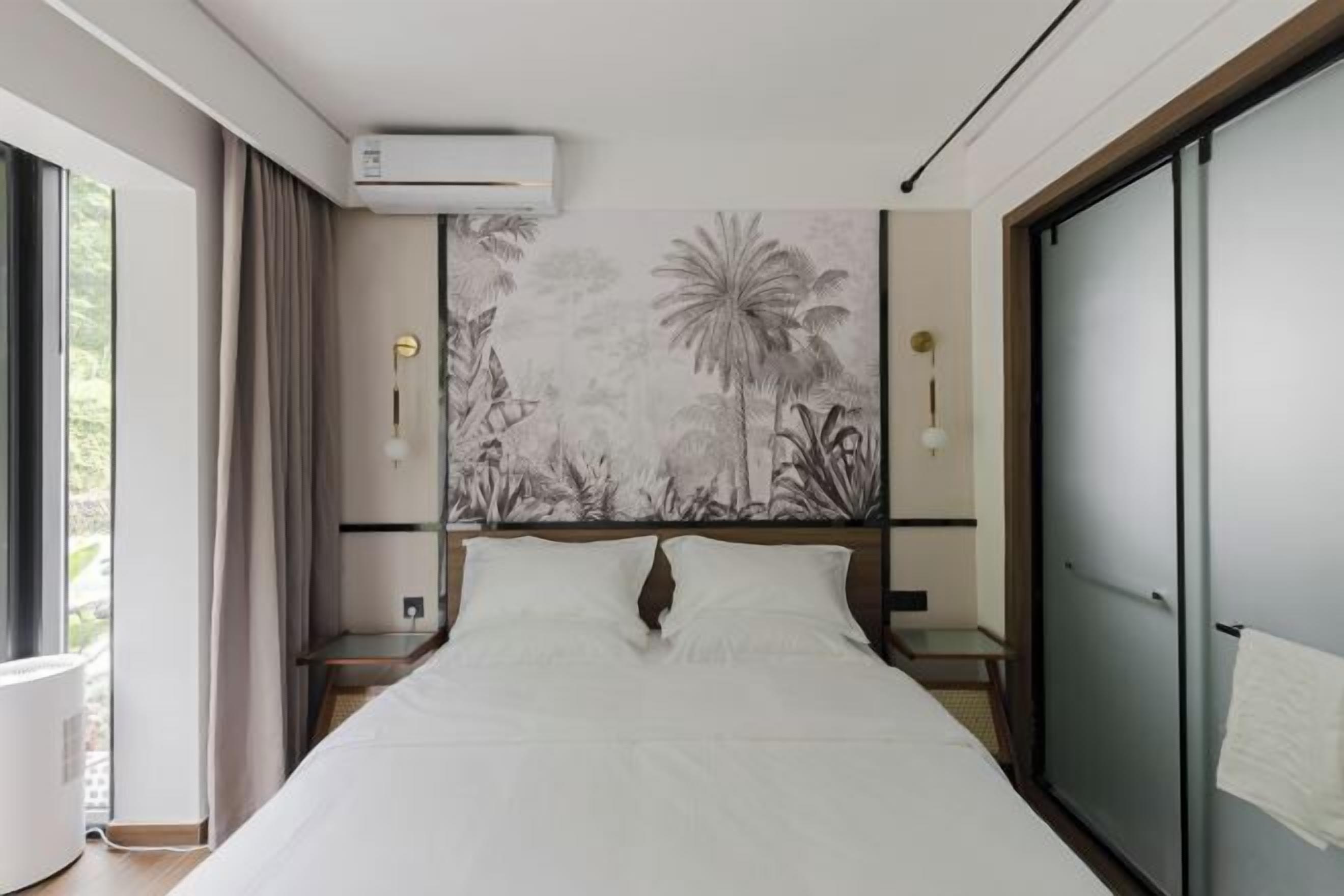 A hotel room with a bed, a wall-mounted air conditioner, a nightstand, a bedside lamp, and a large window.