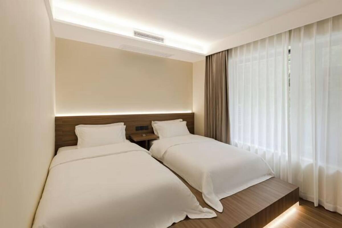 A hotel room with two beds, a wooden headboard, a nightstand, and a window with sheer curtains.