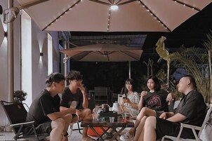 A group of people seated at an outdoor table under a large umbrella, with chairs and a table lamp visible.