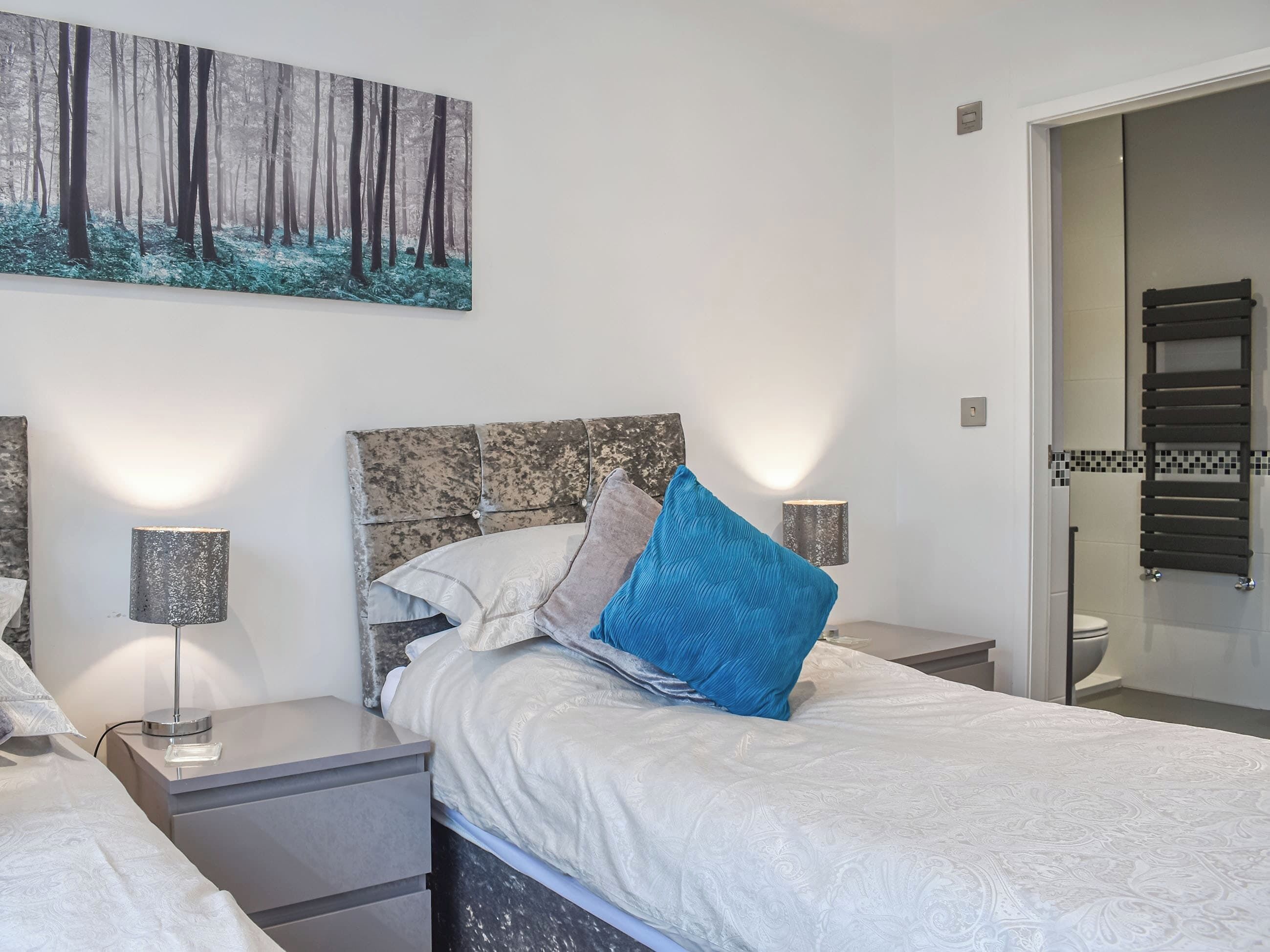 A bedroom with a bed, two bedside tables, a nightstand, a wall-mounted painting, and a bathroom door.