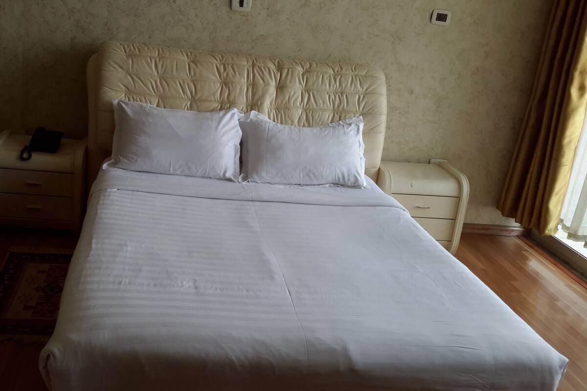 A neatly made bed with white linens and pillows, a bedside table with a lamp, and a window with curtains.