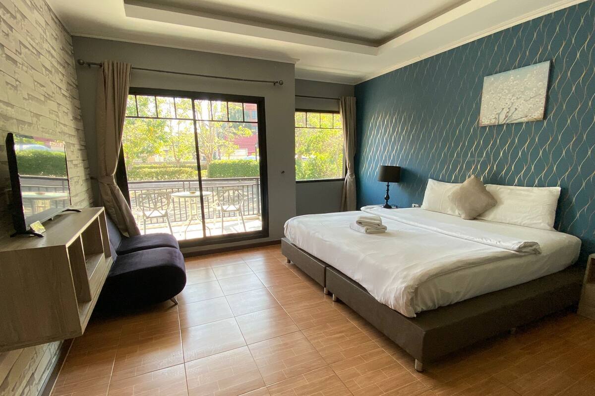 A modern hotel room with a large bed, a TV on a wooden console, a balcony with outdoor furniture, and a blue patterned wall.