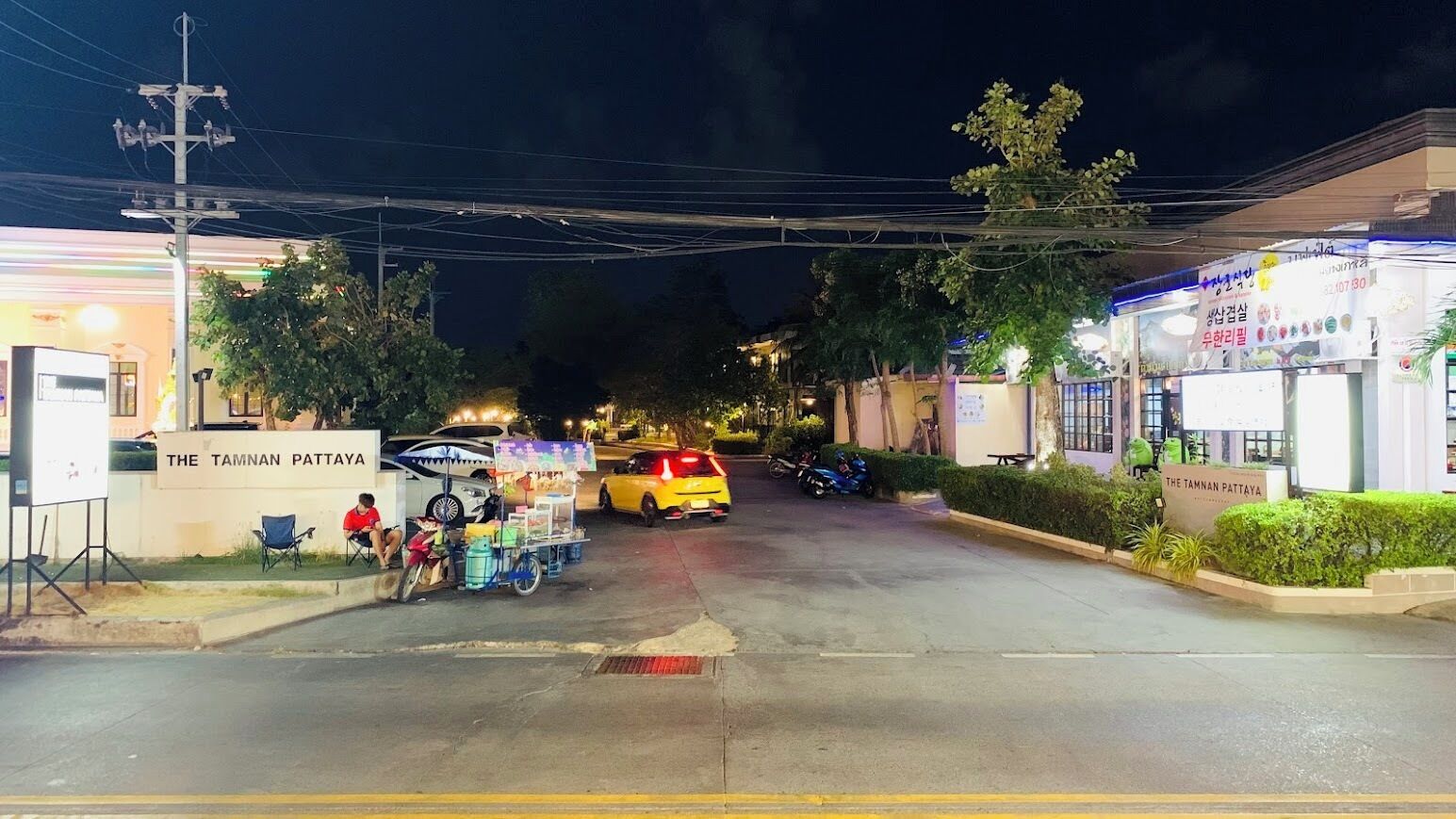 Vista nocturna de una calle con un puesto de comida, coches aparcados y un edificio que dice 'The Tamnan Pattaya'.
