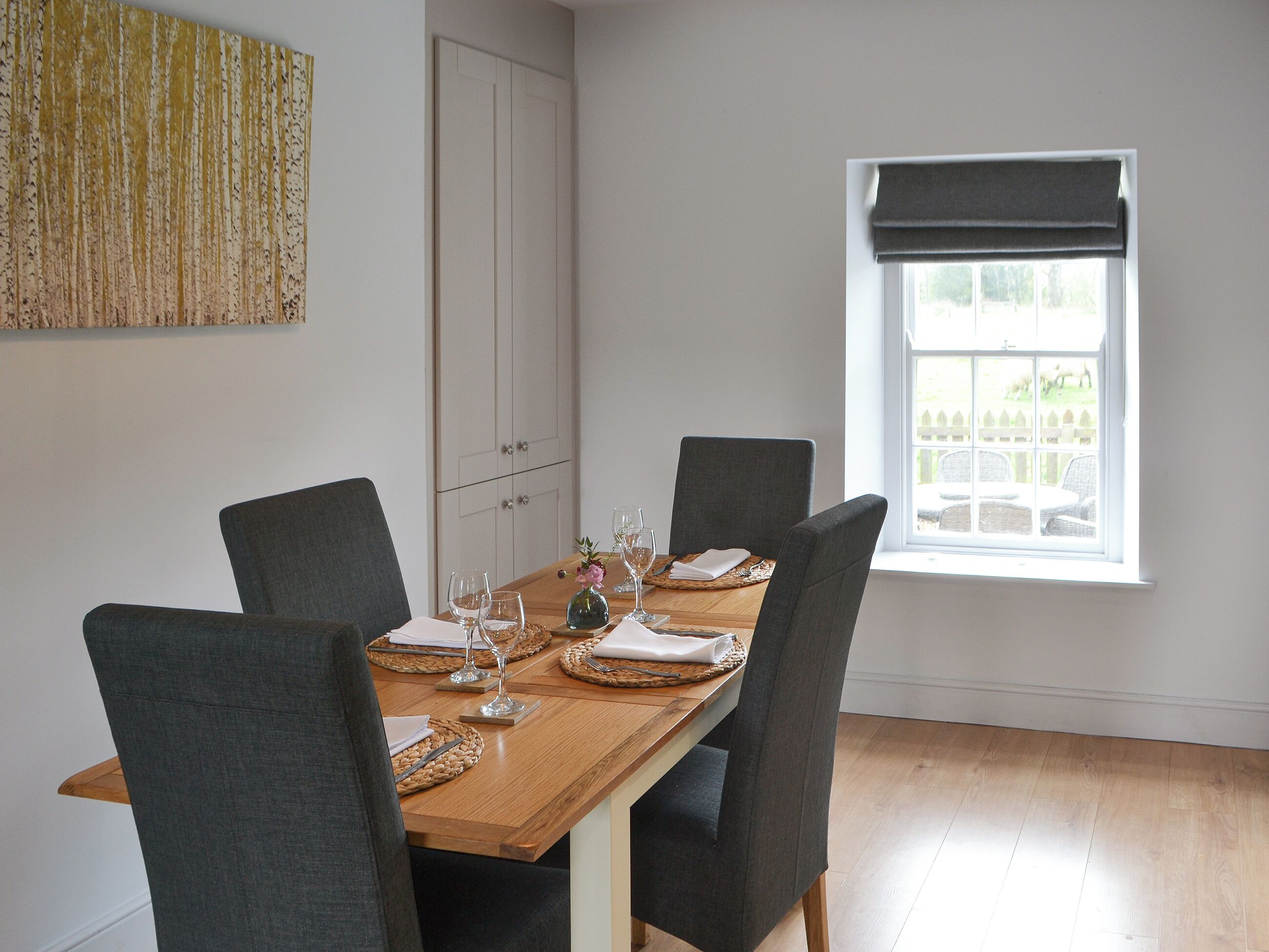 Een eetkamer met een houten tafel voor vier personen, grijs beklede stoelen, een raam met uitzicht op een hek, en een kast met zilveren handgrepen.