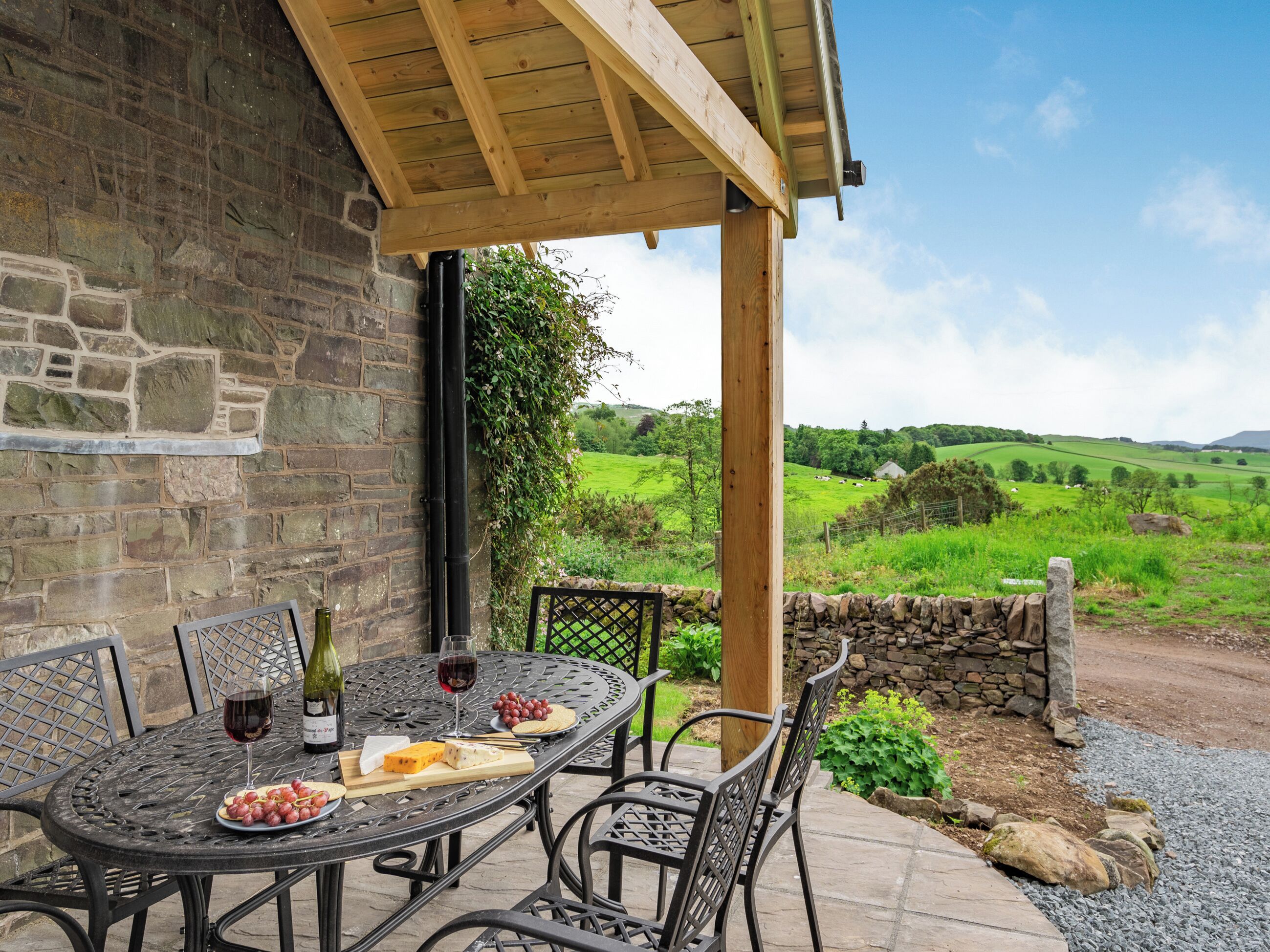 Un bâtiment en pierre surmonté d’une pergola en bois, une table avec une bouteille de vin, du fromage et des raisins, et une vue sur des champs et des collines verdoyants.