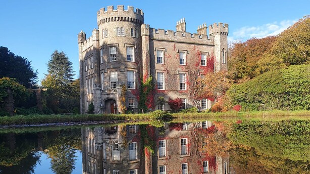 Un bâtiment qui ressemble à un château, avec un fossé, et dont l’image se reflète dans l’eau, ainsi que celle de la structure et des arbres environnants.