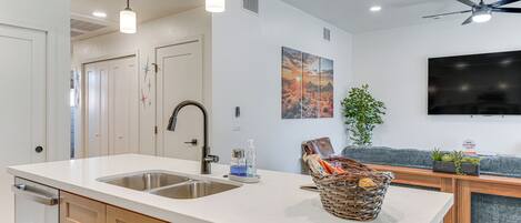 Una cocina moderna con una isla grande, pileta de acero inoxidable y armarios de madera. Cuenta con un ventilador de techo, iluminación empotrada y un televisor de pantalla plana montado en la pared.