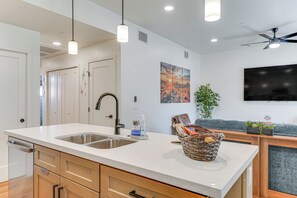 A modern kitchen with a large island, stainless steel sink, and wooden cabinets. There is a ceiling fan, recessed lighting, and a flat-screen TV mounted on the wall. - Newly Finished, High-End Condo Near Tucson’s Best! (Tucson)