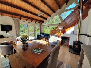 Een ruime woonkamer met houten plafond, een open haard, een bank, een eettafel met een boek en een televisie aan de muur.