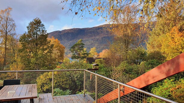 Eine Holzdielen-Terrasse mit Metallgeländer, die auf eine gebirgige Landschaft mit Herbstlaub blickt.