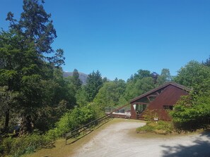 A rustic cabin with a gravel path leading to it, surrounded by dense forest and a mountain in the background. - Arkaig Lodge - UK7254 (Spean Bridge)