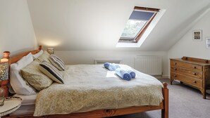 Een slaapkamer met een schuine kap, een dakraam, een houten bed met beddengoed met een patroon, een nachtkastje met een lamp en een dressoir.
