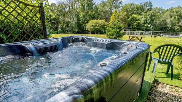 Een hottub in een tuin met een houten terras, omringd door groen en een picknicktafel.