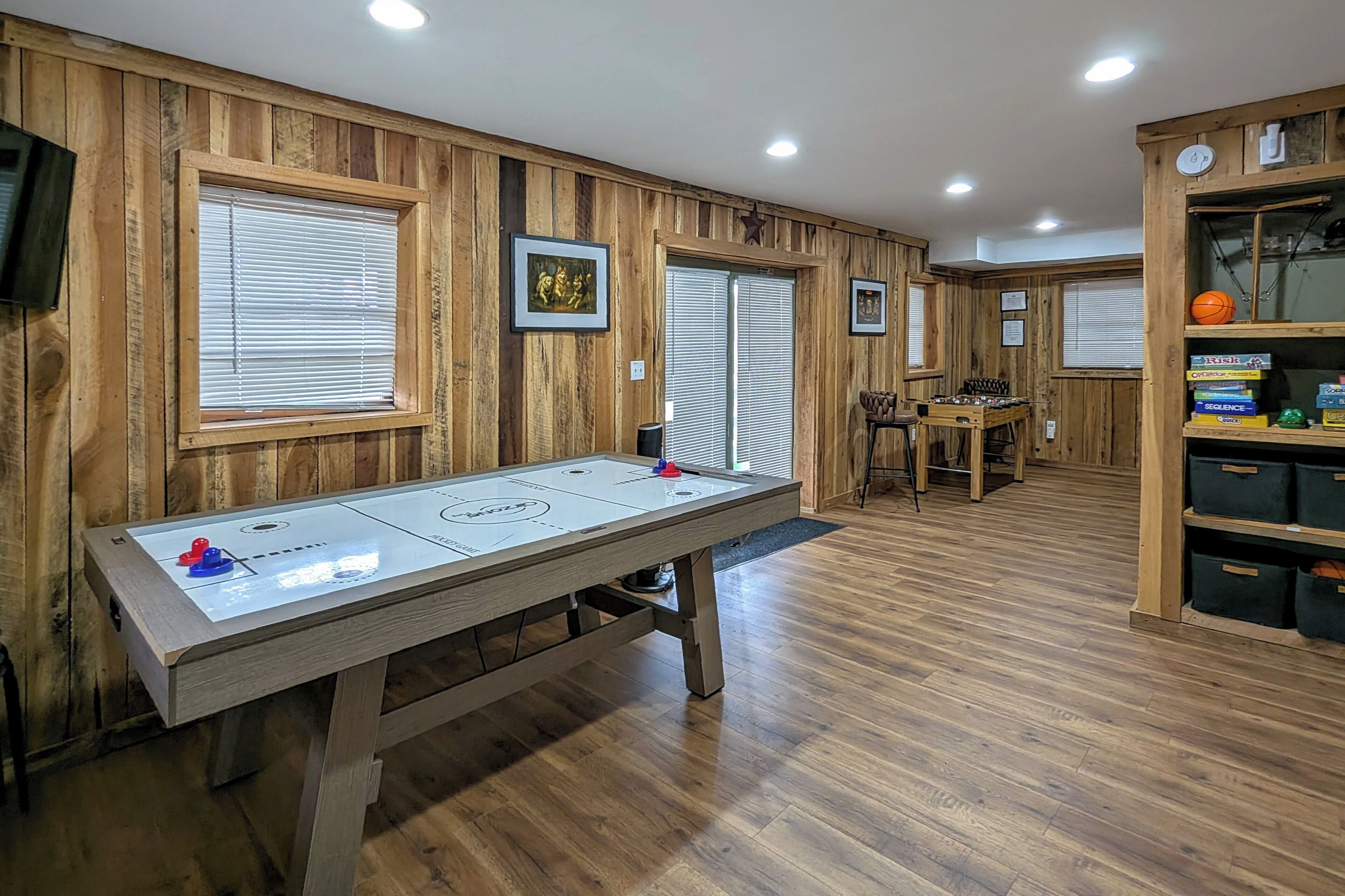A room with wooden paneled walls, a shuffleboard table, a billiards table, and a bookshelf with storage boxes.