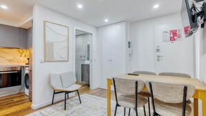 A modern kitchen with a built-in oven and washing machine, a dining area with a wooden table and four chairs, and a bathroom visible through an open door. - Vita Portucale | Alfama Sweet Retreat (Lisboa)
