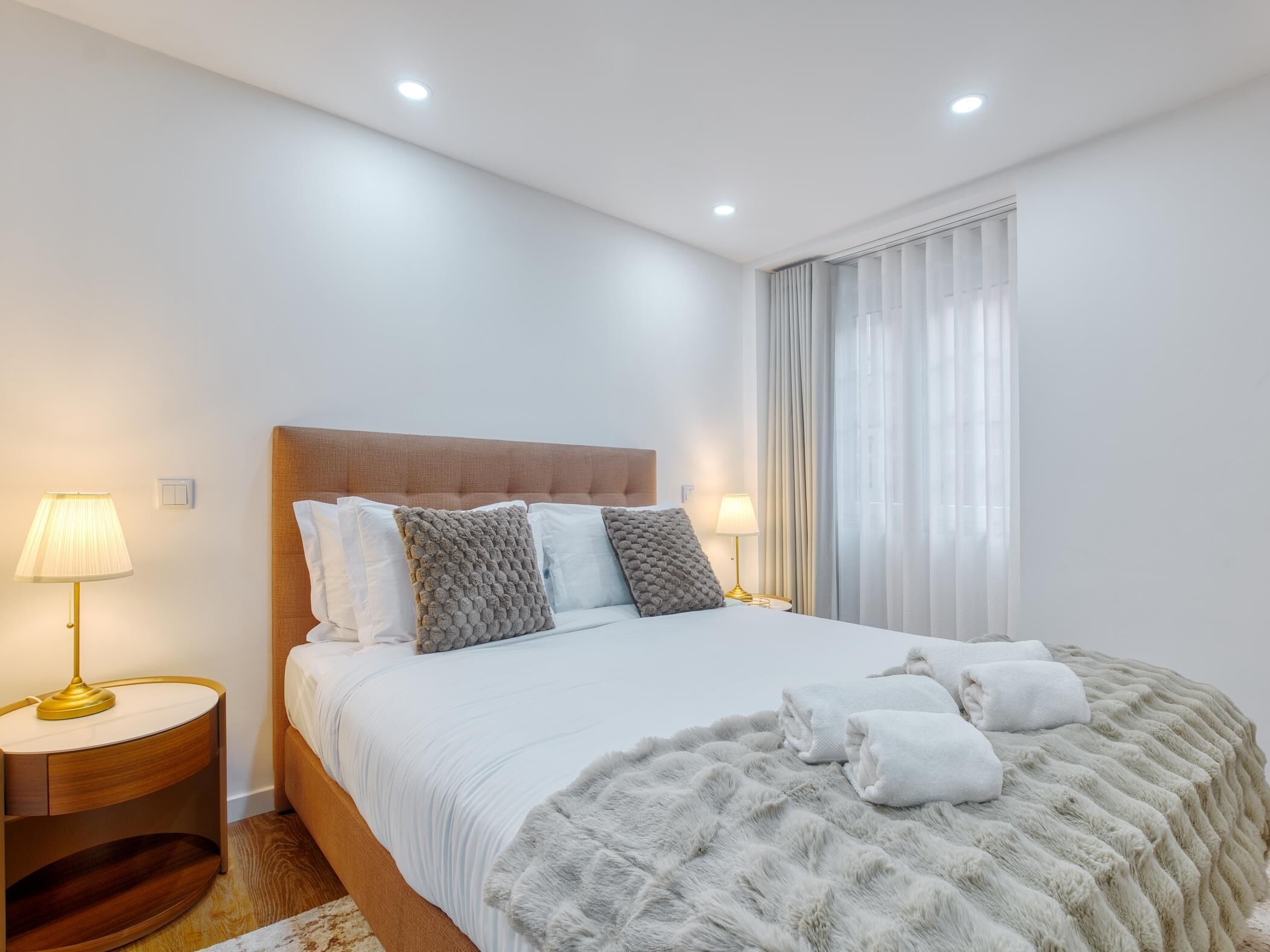 Habitación de hotel moderna con una cama grande, dos mesitas de noche con lámparas, una manta suave y vistas a la ciudad a través de cortinas translúcidas.