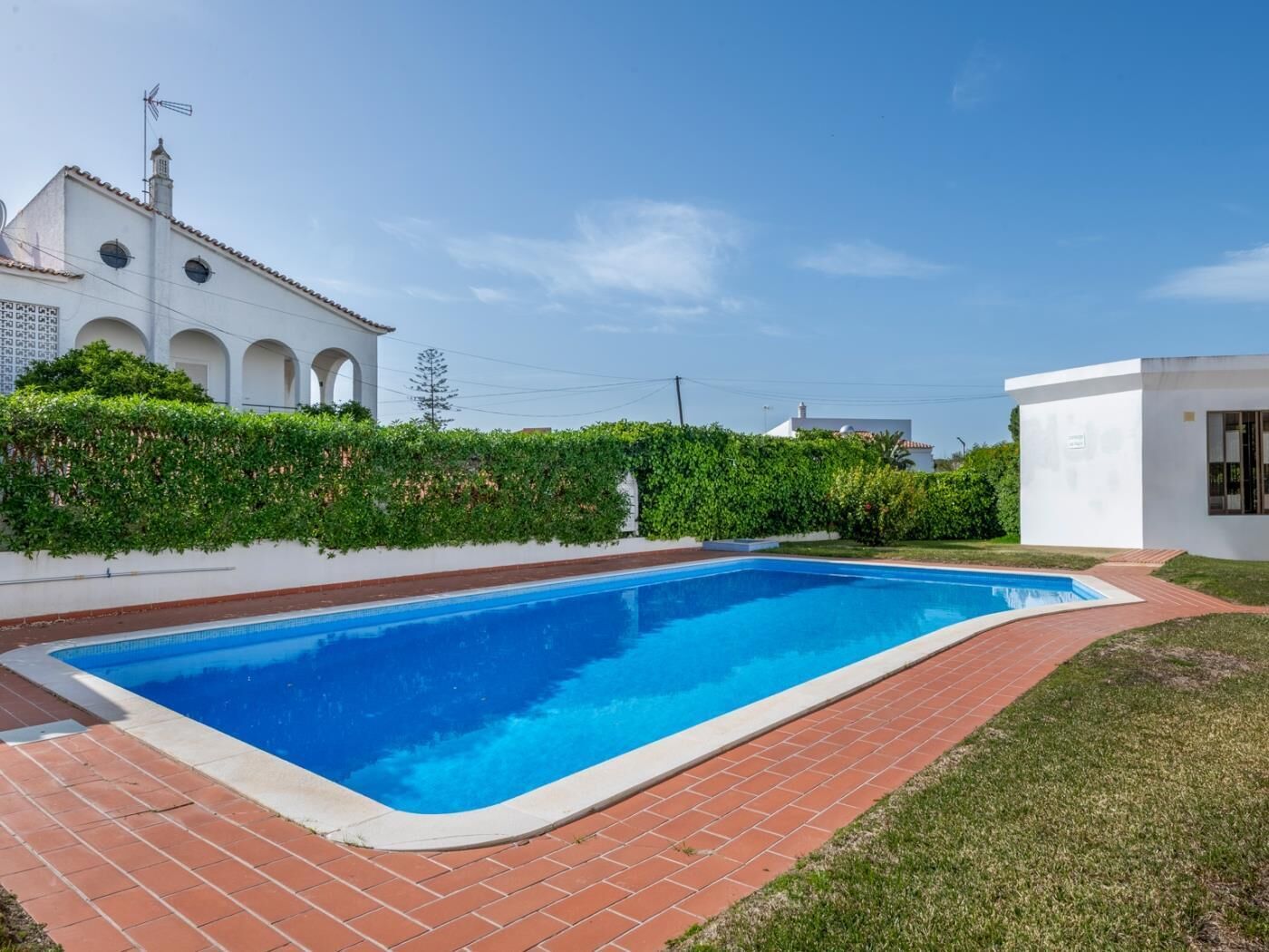 A swimming pool with a brick border, surrounded by a grassy area and a white building with arches.